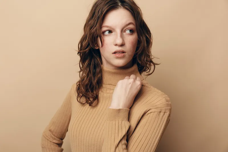 Young woman with curly brown hair wearing beige sweater looks to the side with thoughtful expression. Studio portrait on neutral background. Concept of contemplation and casual fashion.