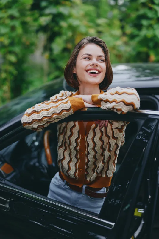 Joyful woman leans on a car door, smiling brightly in a sunny outdoor setting, captured in a candid lifestyle moment.