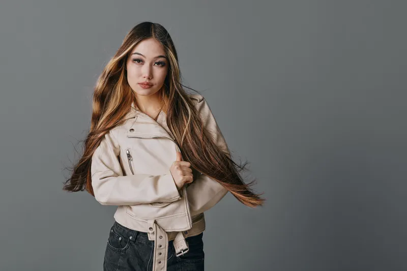 Young woman with long brown hair wearing light jacket and jeans standing against gray background looking at camera with confident expression and natural makeup.