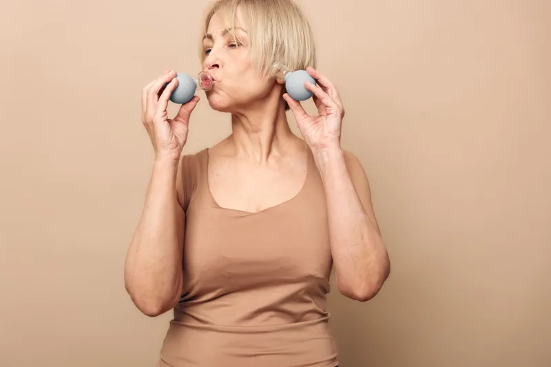 Mature woman with short blonde hair holds two grey massage balls near her face, purses lips as if blowing air. Neutral background and beige clothing create calm wellness concept.