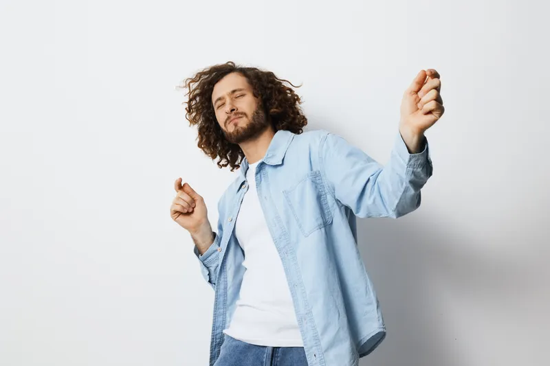 Young man with curly hair dancing joyfully in casual attire against a minimalist white background, capturing the essence of happiness and carefree style