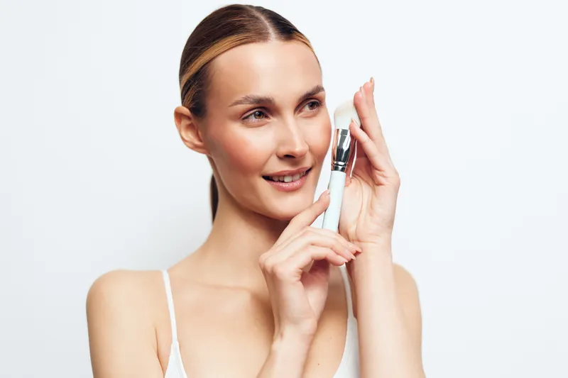 Young woman with blonde hair gently holds a white cosmetic bottle near her face while smiling softly against a plain white background, showcasing skincare routine and beauty care.