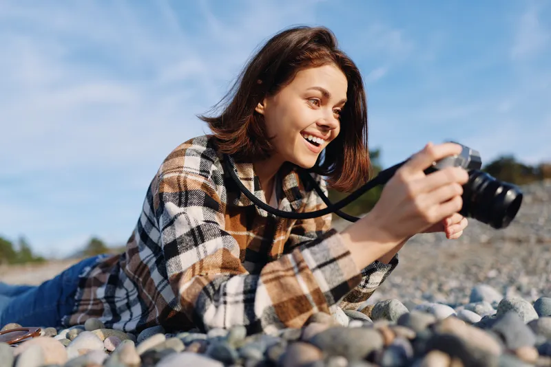 Woman photographer lying on a pebbled shore, smiling as she captures with a camera, outdoors scene with sunlight, casual style and candid energy, showing hobby photography and nature in a relaxed