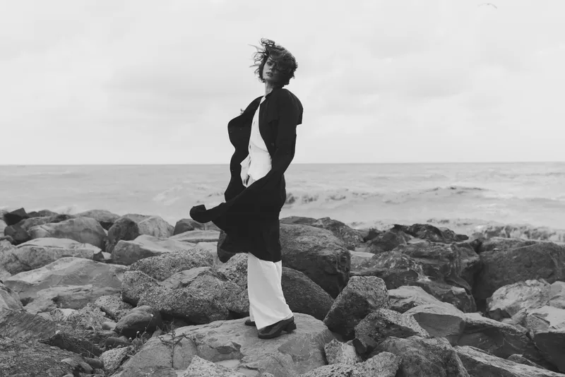 Woman on rocky shore facing the sea, wind lifting her dress in a solitary landscape, black and white mood, dramatic composition, serene motion on a coastal scene suitable for fashion and nature