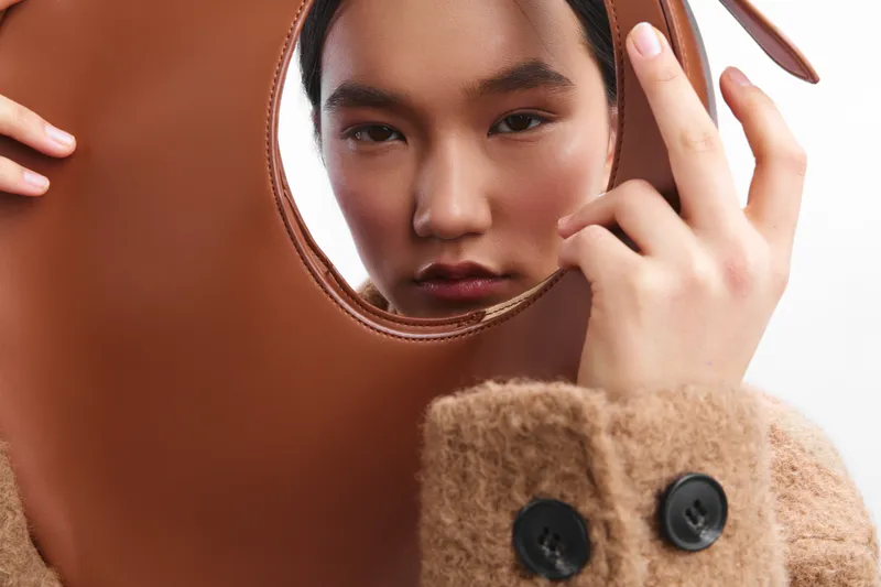 asian woman peers through a circular cutout in a stylish leather bag, presenting a poised, confident look that blends fashion accessory concepts with contemporary street style photography.