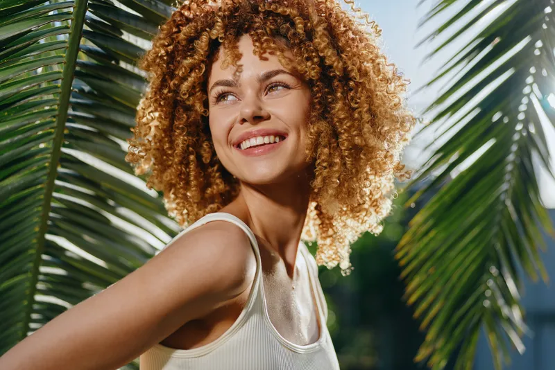 smiling woman with curly hair stands near tropical palm leaves in sunlight, wearing white tank top, radiating happiness and natural beauty outdoors