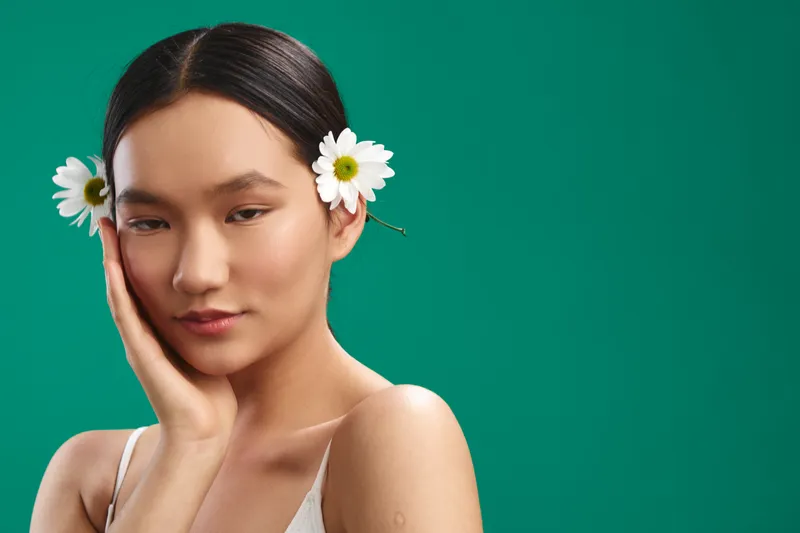 asian woman with lightweight makeup poses against a vibrant green backdrop, wearing a simple white outfit, flowers tucked in her hair, creating a calm and elegant studio portrait.