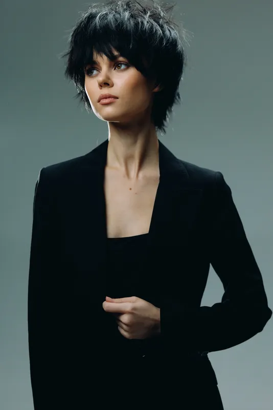 Stylish woman with short black hair wearing a black blazer, posing confidently against a neutral gray background in a minimalistic style.