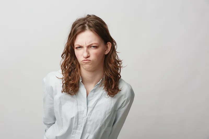 Young woman with curly hair wearing white shirt shows displeased facial expression, frowning and looking at camera. Isolated on light grey background. Concept of dissatisfaction and emotion.