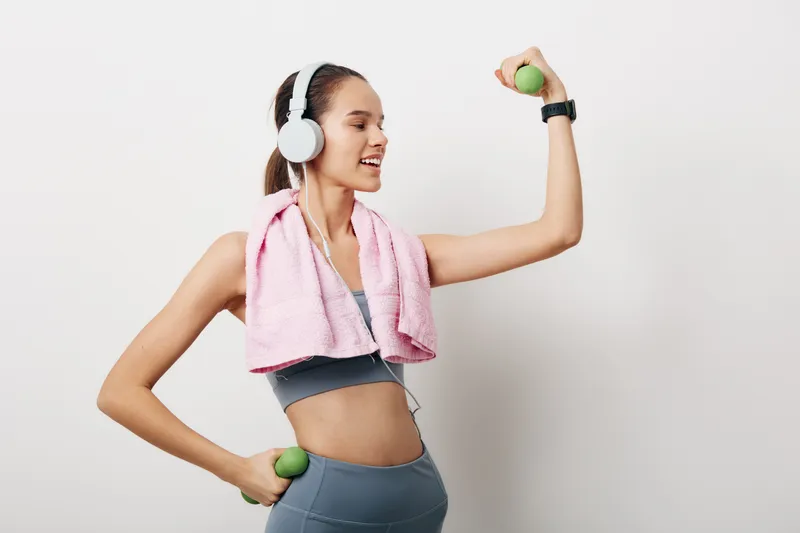 Woman fitness sport portrait exercising with small green dumbbells, wearing sports bra and leggings, pink towel around neck, white headphones and smartwatch on plain white background.