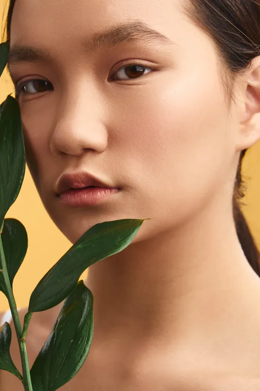 Asian woman is featured in a closeup portrait with green plant leaves softly framing the face, conveying natural beauty, calm confidence, and contemporary elegance.