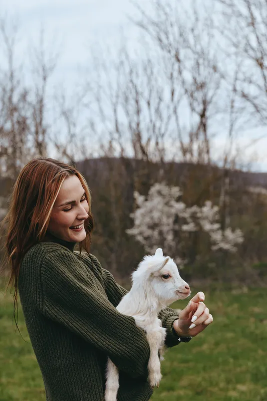 Cheerful woman outdoors holds a white goat kid in a green field, showcasing a warm lifestyle moment, natural bond, peaceful countryside portrait, and simple joy in nature, with soft sunlight
