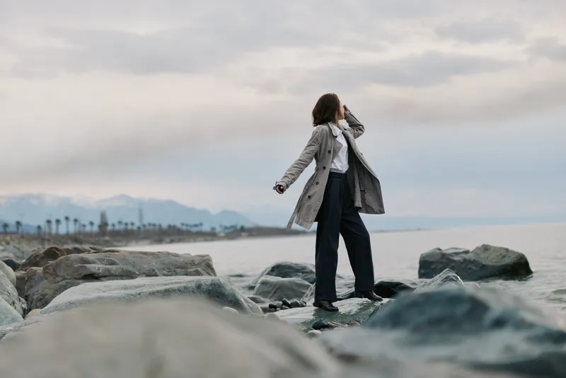 Woman in trench coat stands on rocks near sea shore looking into distance. Cloudy sky and calm water create peaceful atmosphere. Concept for travel, exploration, and outdoor lifestyle.