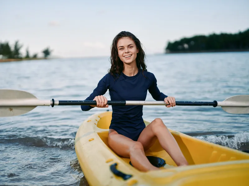 Smiling young woman paddles a yellow kayak across calm water, wearing a navy swimsuit, with a distant shoreline and soft clouds in the backdrop.