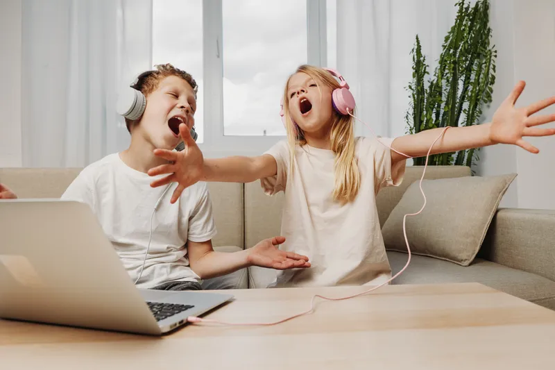 Two happy children wearing headphones sing passionately together at home, enjoying music with a laptop on the table in a bright living room setting.