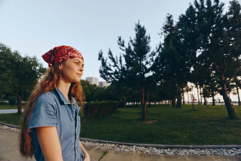Young woman with red bandana walking outdoors in park during late afternoon, casual denim outfit, natural light, green trees and urban background.