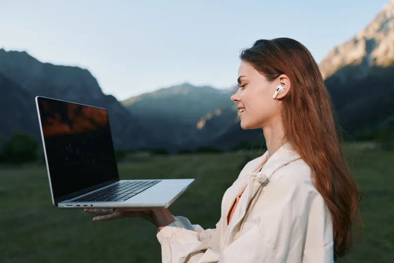 A cheerful professional woman holds a laptop outdoors, smiling and focused, wearing wireless earbuds in a scenic mountain landscape, conveying connectivity and remote work.