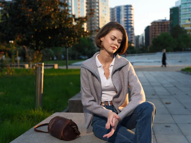 A calm urban professional woman sits on a bench in a modern city park, looking thoughtful and relaxed as sunlight creates a warm, reflective atmosphere.