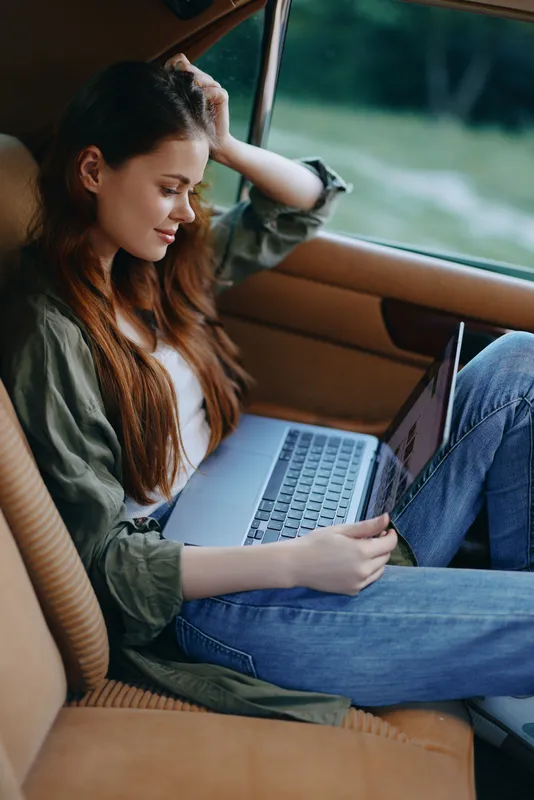 Young woman with long hair working on a laptop in a car, wearing casual clothing, showcasing a relaxed and focused expression in a natural outdoor setting.