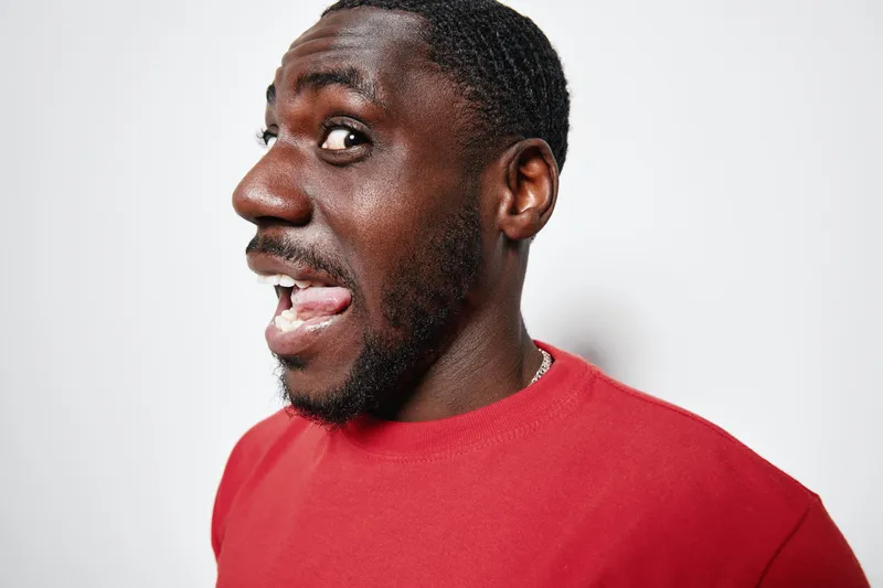 African American man with dark skin wearing red shirt making funny face with tongue out and eyes wide open against white background. Portrait, expression and casual style.