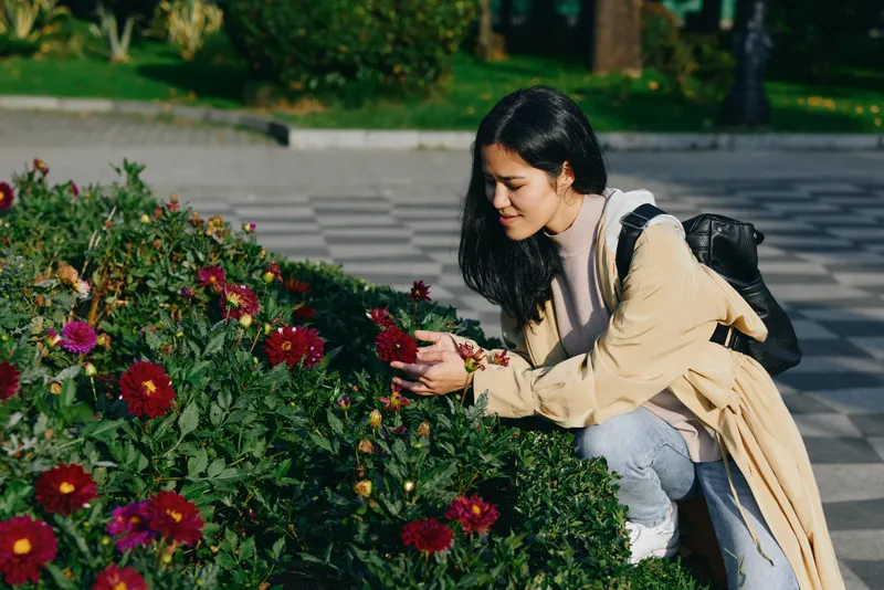 Woman crouching beside vibrant flowers in a park, admiring blossoms in an outdoor scene, casual attire, backpack ready for a stroll, nature inspired moment for leisure.
