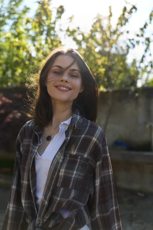 A smiling woman in a plaid shirt stands outdoors in daylight, capturing a casual lifestyle moment with natural confidence, relaxed posture and bright surroundings