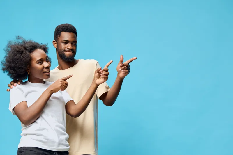 Young African couple joyfully pointing at each other against a vibrant blue background, expressing happiness and connection in a playful moment