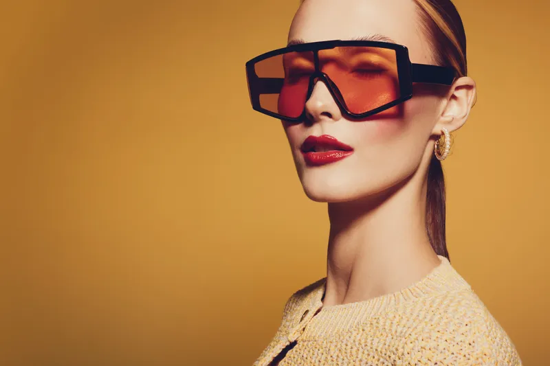 Stylish woman with oversized orange sunglasses and red lipstick poses against yellow background. Fashion portrait with modern accessories and confident expression in studio lighting.