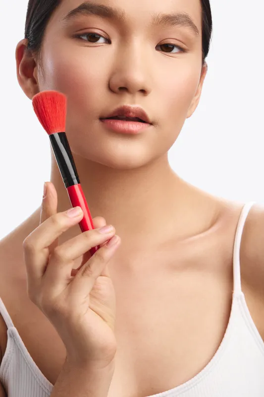 asian woman is featured in a studio portrait as she uses a makeup brush, showcasing smooth skin and soft lighting with a clean background for a beauty focused scene.