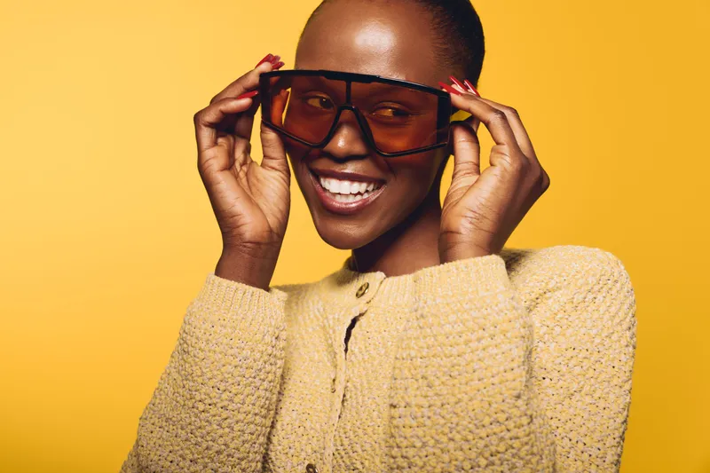 Smiling dark skinned African woman with short hair adjusts large sunglasses, wearing beige knitted sweater. Studio portrait on yellow background. Concept of fashion, beauty, and confidence.