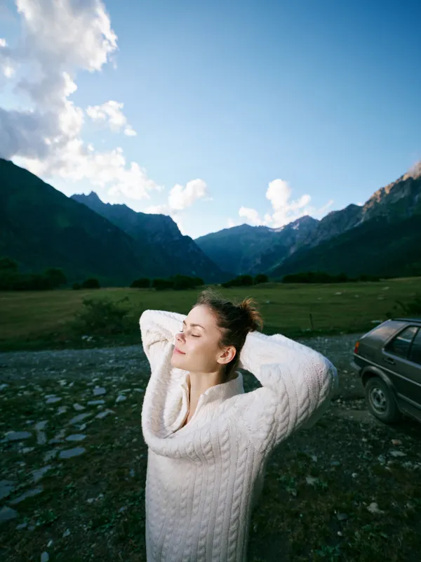 Mountain valley scenery with a relaxed woman in a cozy knit sweater, arms behind her head, enjoying fresh air and sunlit nature in a wide open countryside on a bright day