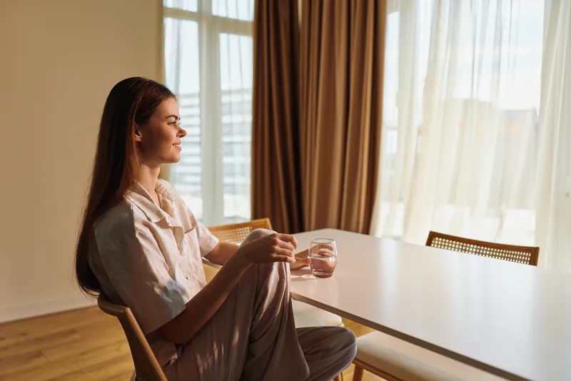Woman portrait relaxation morning home sunlight: a female sitting at a dining table holding a glass, gazing through window in warm natural light, calm cozy interior and thoughtful quiet moment.