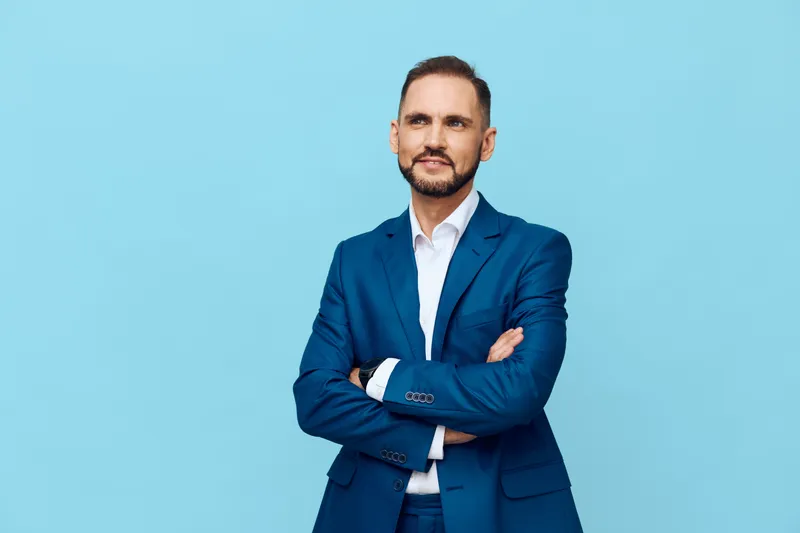 A confident business professional stands with arms crossed against a solid blue background, expressing calm focus, pride, and readiness for success.