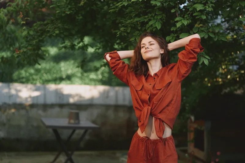 Young woman enjoys outdoor lifestyle, wearing rust orange outfit, stands with arms raised, relaxed joyful mood, natural garden backdrop, sunlit scene, candid fashion portrait