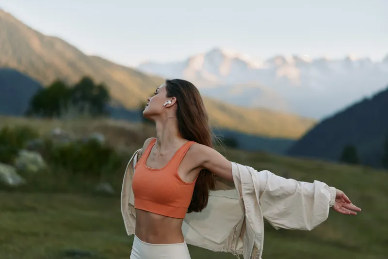 A woman wearing an orange sports top and a light jacket stands outdoors in an alpine landscape. She stretches her arms wide, face lifted, smiling, enjoying fresh air and nature tranquil energy.