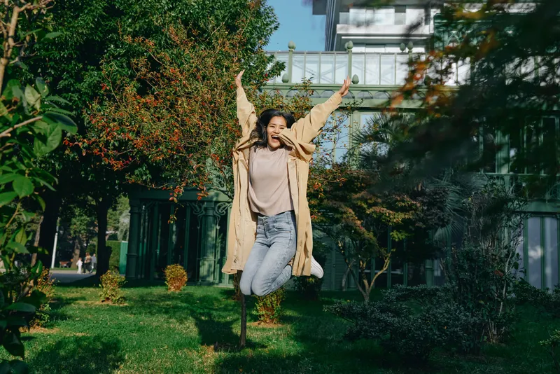 Joyful jump outdoors by greenery and a modern building, casual coat, sunny day, carefree moment, lifestyle scene, energetic movement, urban park setting, vibrant atmosphere