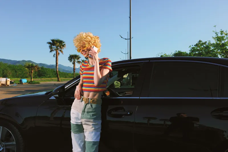 A woman wearing a curly blond wig in a striped top leans on a sleek car in a sunny parking lot, capturing retro film color and casual street fashion. Concept focuses on bold style and playful mood.