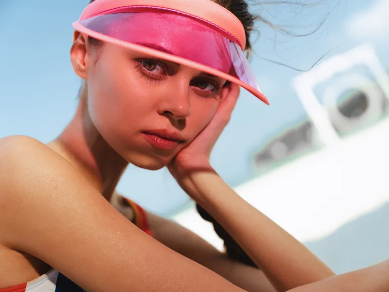 Portrait of a young woman wearing a pink visor on a sunny beach, close up, sunlit skin, thoughtful expression, vibrant colors, summer vibe and carefree mood.