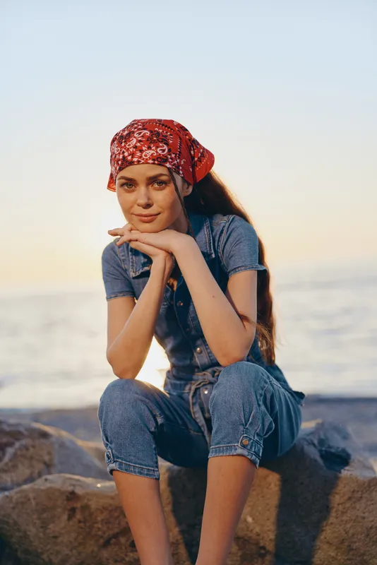 Young woman wearing red bandana and denim outfit sits on rocks by the sea during golden hour, smiling softly with hands under chin in relaxed pose.