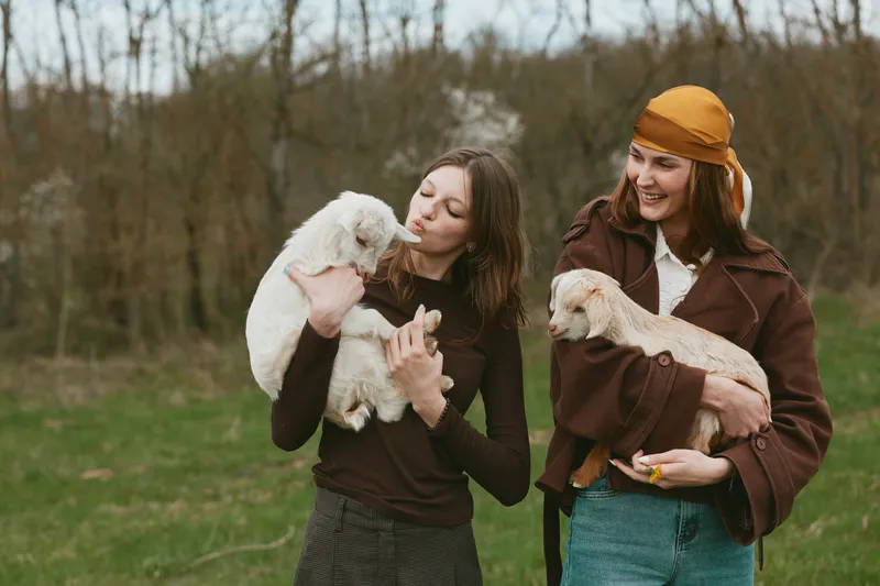 Two women outdoors enjoying a playful moment with fluffy puppies, casual autumn outfits, soft natural light, grass and trees create a warm lifestyle mood with friendship