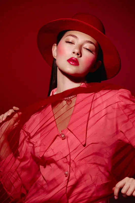 Young woman in red hat and sheer dress poses with closed eyes against red backdrop. Fashion and beauty creative portrait with strong stylish mood and dramatic shadows.