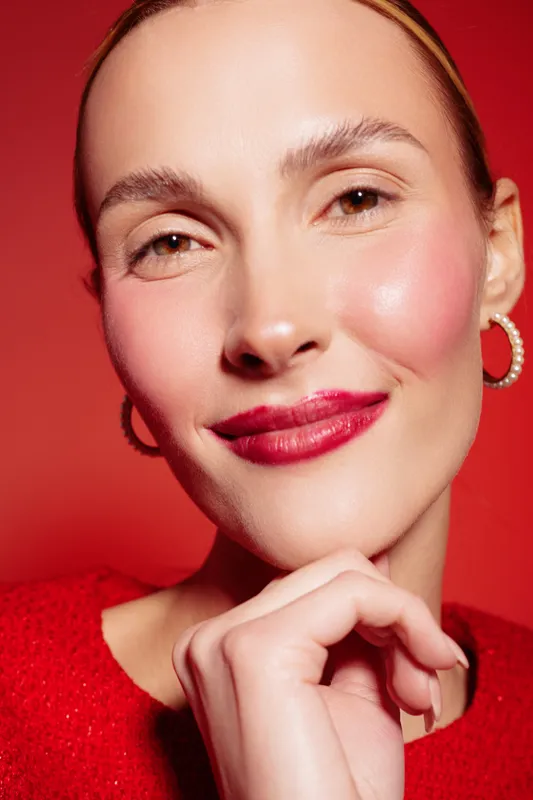Close-up portrait of a smiling woman with red lipstick and pearl earrings on red background. Concept of beauty, fashion, makeup, and confident female model with natural skin glow.