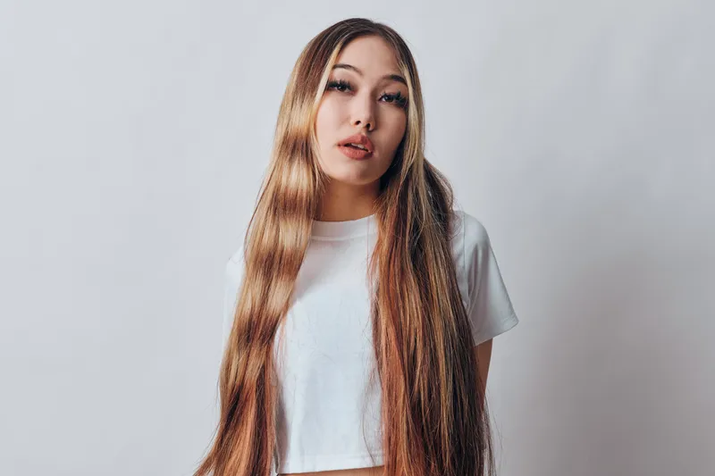 Young woman with long straight hair wearing white crop top posing in studio with neutral expression. Asian female model with natural makeup and relaxed casual style on plain background.