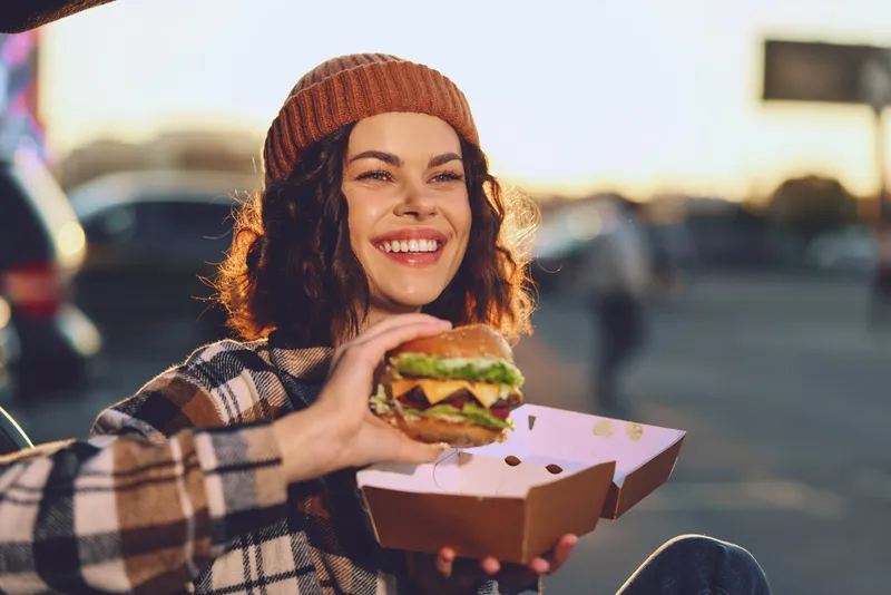 Woman burger smile authenticity candid lifestyle golden glow: young woman outdoors in beanie holding a takeaway burger in a box, laughing in warm sunset light with emotional storytelling mood.