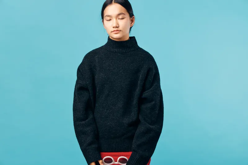 Young woman with braided hair wearing a black sweater stands with eyes closed against blue background. Minimalist portrait with calm expression and casual style in studio lighting.