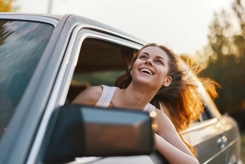 Young woman smiling joyfully while leaning out of a car window, enjoying a sunny day, representing freedom and adventure.