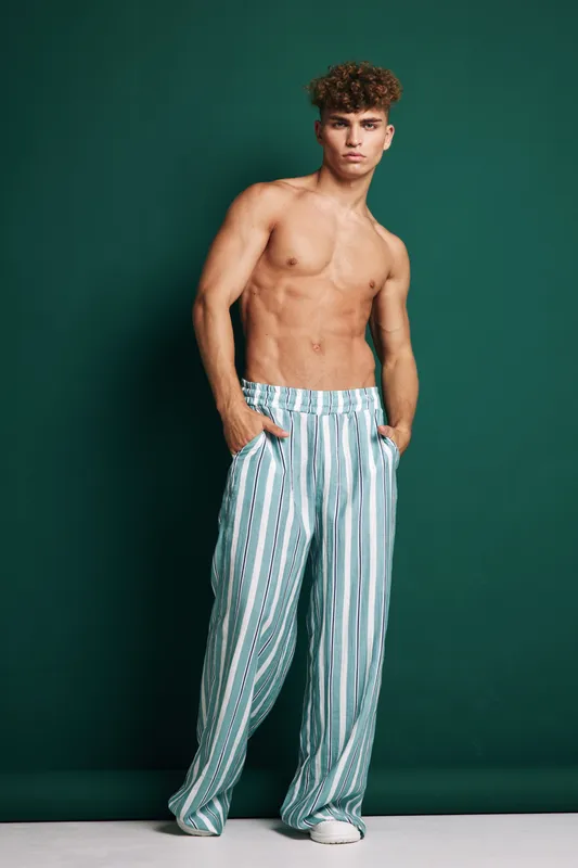 Young man with curly hair posing shirtless in striped wide pants and white sneakers against green background. Fashion model standing with hands in pockets and confident expression.