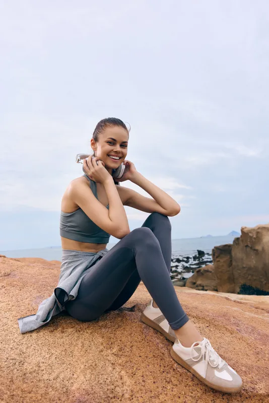 Young female athlete in activewear enjoying music on a rock, showcasing a healthy lifestyle and happiness, with a serene landscape background
