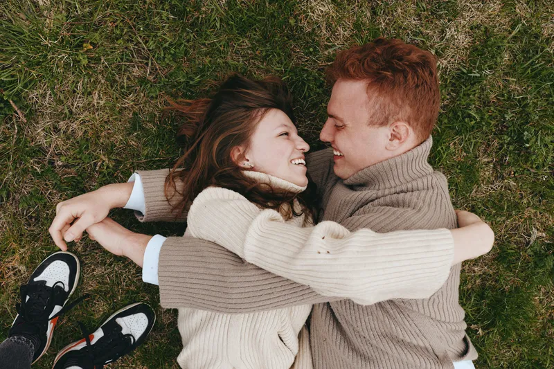 Cheerful couple outdoor on grass, embracing, smiling with eyes closed, sunlit moment, cozy knitwear, relaxed posture, natural colors, intimate connection, candid lifestyle moment, affection, happiness