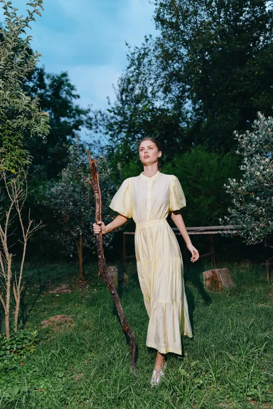 Woman in a pale yellow dress stands in a lush garden holding a tall staff, exuding calm confidence and ethereal natural beauty in a bright outdoor setting